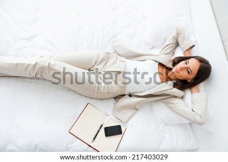 Relaxing in hotel room. Top view of beautiful young businesswoman in suit holding hands behind head and keeping  while lying in bed at the hotel room