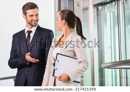 Discussing project with colleague. Two cheerful business people discussing something and smiling while getting out from elevator