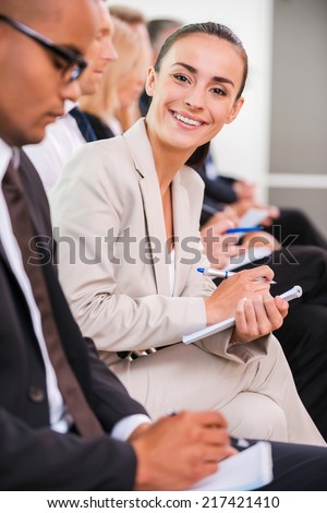 Businesswoman at the conference. Side view of business people sitting in a row and writing something in their note pads while confident young woman looking at camera and smiling