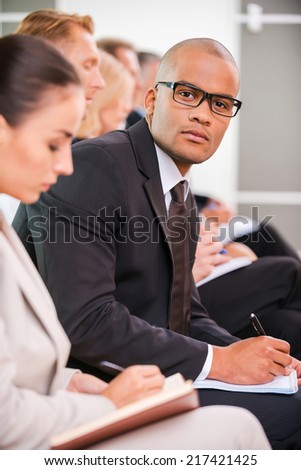 Businessman at the conference. Side view of business people sitting in a row and writing something in their note pads while confident African man in eyewear looking at camera