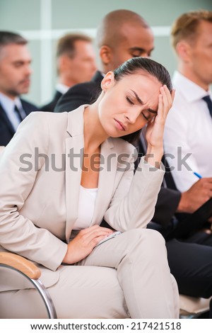 Suffering from awful headache. Depressed young man holding head in hand and keeping eyes closed while sitting at the chair in conference hall with people in the background