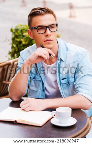 Waiting for inspiration. Thoughtful young man touching his chin with pen and looking away while sitting in sidewalk cafÃ?Â?Ã?Â© with note pad on the table