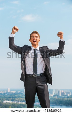 I did it! Happy young man in formalwear keeping arms raised and expressing positivity while standing outdoors with cityscape in the background
