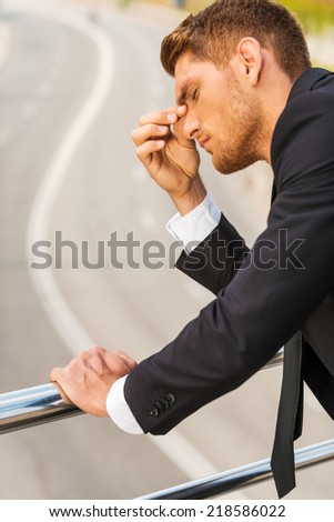 Depressed businessman. Depressed young man in formalwear touching his nose and keeping eyes closed while standing outdoors and leaning at the metal railing