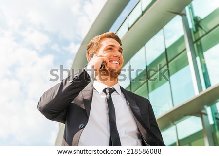 Confident business talk. Cheerful young man in formalwear talking on the mobile phone and looking away while standing outdoors and against building structure