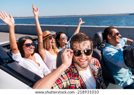 Summer road trip. Group of young happy people enjoying road trip in their convertible while handsome man adjusting his sunglasses and smiling