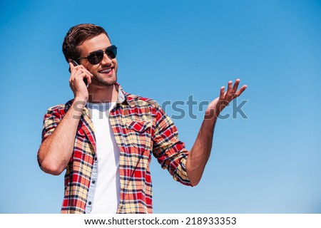 Carefree talk. Cheerful young man in sunglasses talking on mobile phone and gesturing while standing outdoors with blue sky as background
