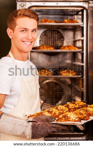 Fresh baked croissant for you. Handsome young man in apron taking the fresh baked croissants from the oven and smiling