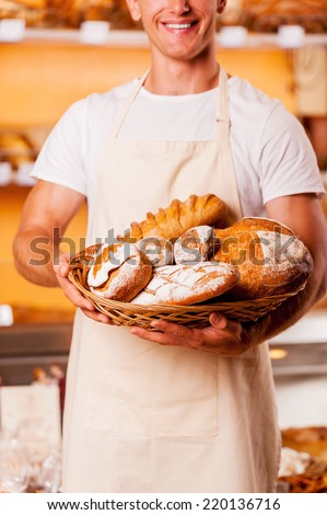 The best baked goods in town. Cropped image of young man in apron holding basket with baked goods and smiling while standing in bakery shop