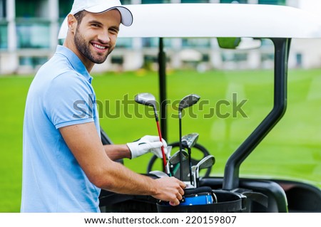 Choosing the proper driver. Handsome young male golfer choosing driver while standing near the golf cart