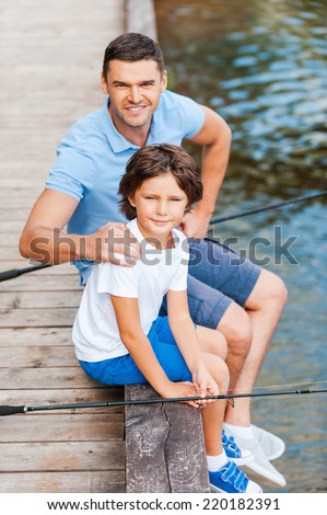 Spending great time together. Top view of father and son looking at camera and smiling while sitting at the quayside with fishing rods laying near them