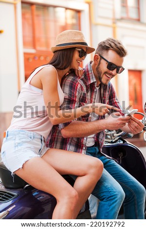 Look at that! Beautiful young couple sitting on scooter together while happy woman showing something at mobile phone