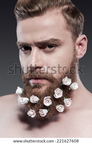 Taking good care of his beard. Portrait of handsome young shirtless man with flowers in his beard looking at camera while standing against grey background