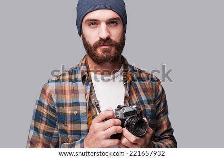 Confident photographer. Handsome young bearded man holding old-fashioned camera and looking at camera while standing against grey background