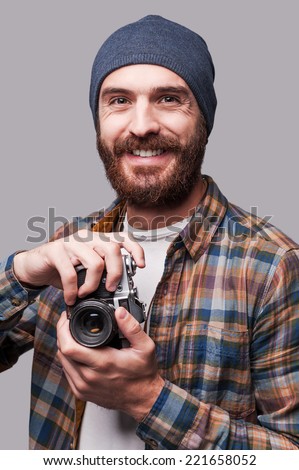Confident photographer. Handsome young bearded man holding old-fashioned camera and smiling while standing against grey background