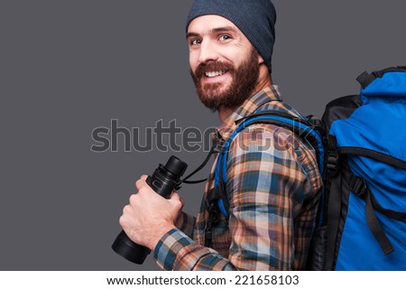 Confident tourist. Side view of handsome young bearded man with backpack holding binoculars and smiling while standing against grey background