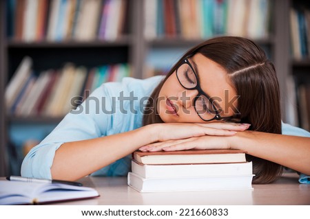 Exam exhaustion. Tired young women holding her head on the book stack and sleeping while sitting at the library desk
