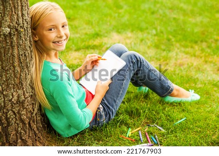 Little artist. Rear view of cute little blond hair girl drawing something at her note pad and looking over shoulder with smile while sitting on green grass and leaning at the tree