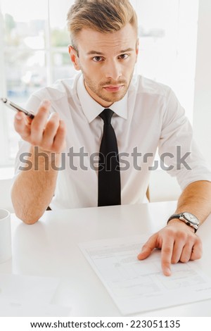 You are fired! Portrait of serious young man in formalwear stretching out a pen and pointing to the paper  while sitting at his work place