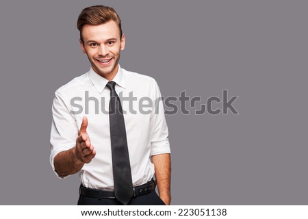 You are welcome in our company! Portrait of cheerful young man in formalwear stretching out hand for shaking while standing against grey background