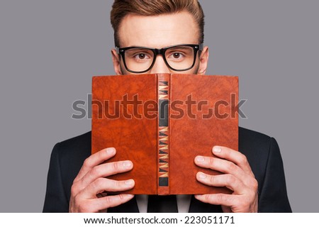 More knowledge!  Young man in formalwear hiding his face behind a book while standing against grey background
