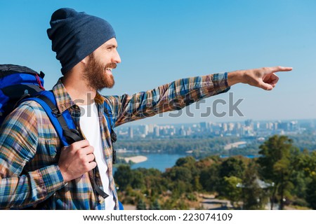 Appreciating the view. Side view of handsome young man carrying backpack pointing and looking away with smile