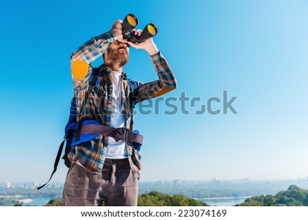 Admiring the majestic view. Handsome young bearded man carrying backpack and looking through binoculars away