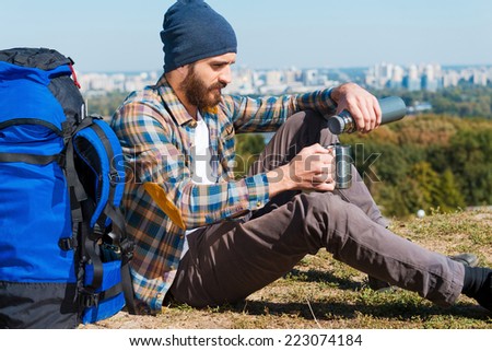 Finally I can relax.  Handsome young man sitting near backpack and pouring tea into a cup