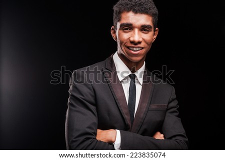 Clothed in confidence. Smiling young Afro-American man keeping arms crossed while standing against black background