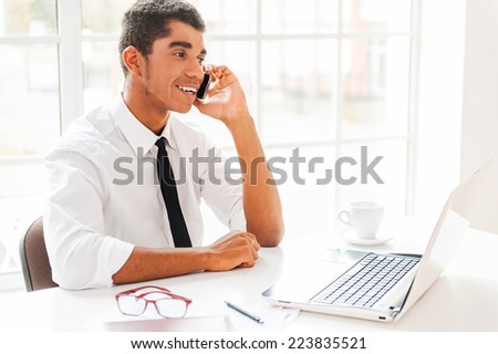 Happy with business going. Happy young Afro-American man in formalwear looking at computer monitor and talking on the mobile phone while sitting at his working place