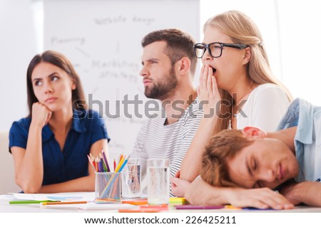 Boring presentation. Group of young business people in smart casual wear looking bored while sitting together at the table and looking away