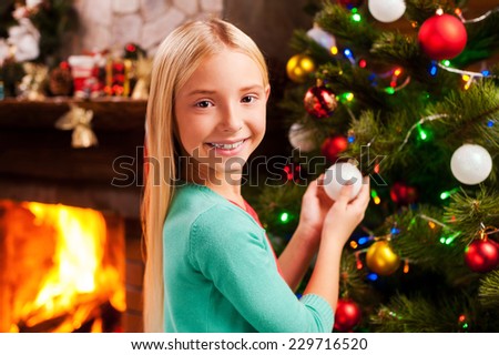 Preparing to Christmas. Cute little girl decorating Christmas Tree and smiling with fireplace in the background
