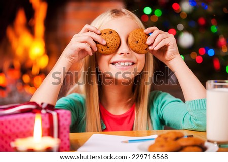 Christmas fun. Cute little girl covering her eyes with cookies and smiling while sitting at the table with Christmas Tree and fireplace in the background