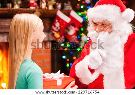 You did not see me. Happy little girl holding gift box and looking at real Santa Claus gesturing silence sign with Christmas Tree and fireplace in the background