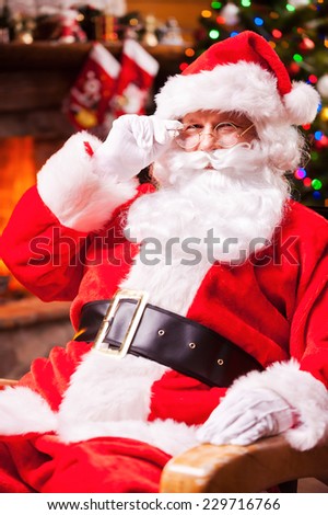 Merry Christmas! Traditional Santa Claus sitting at his chair and adjusting his eyeglasses with fireplace and Christmas Tree in the background