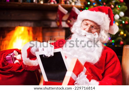 The best present! Cheerful Santa Claus putting a digital tablet into the gift box and smiling while sitting at his chair with Christmas Tree in the background