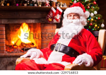 Ho Ho Ho! Traditional Santa Claus sitting at his chair and smiling with fireplace and Christmas Tree in the background