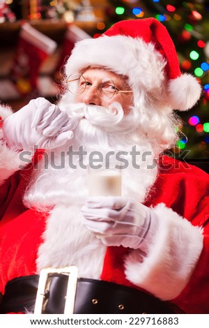 I love milk! Cheerful Santa Claus holding glass with milk and adjusting his mustache with Christmas Tree in the background