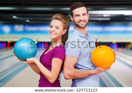 We are a team. Cheerful young man and women looking over shoulders and holding bowling balls while standing against bowling alleys