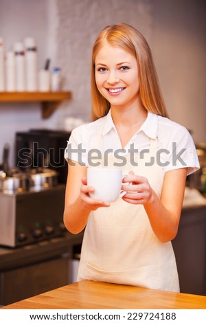 Here you go! Beautiful young female barista stretching out cup of fresh coffee and smiling while standing at the bar counter