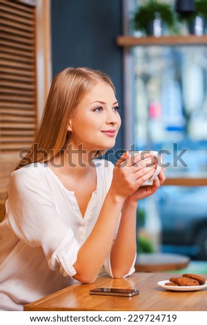 Daydreaming with cup of fresh coffee. Side view of beautiful young smiling woman enjoying coffee in cafe