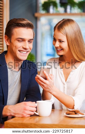 Look at this photo! Beautiful young woman showing something on mobile phone to her boyfriend while enjoying coffee in cafe together