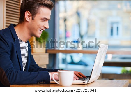 Surfing the net in cafe. Side view of handsome young man working on laptop and smiling while enjoying coffee in cafe