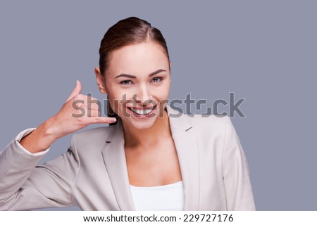 Waiting for your call! Cheerful young businesswoman gesturing phone sign and smiling while standing against grey background