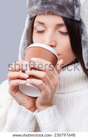 Enjoying hot drink. Close-up of young woman in winter clothing and drinking coffee while standing against grey background