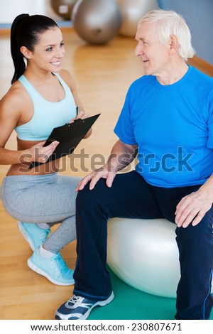 You are making great progress. Confident personal coach holding clipboard and smiling while sitting close to senior man