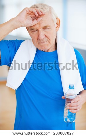 Feeling tired after sports training. Tired senior man with towel on shoulders keeping eyes closed and touching forehead while standing in health club