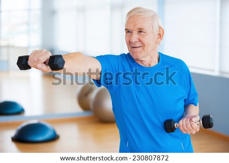 Struggling with age. Confident senior man exercising with dumbbells and smiling while standing in health club