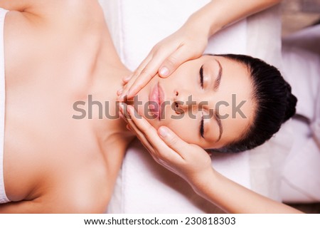 Releasing stress. Top view of beautiful young woman lying on back while massage therapist massaging her face