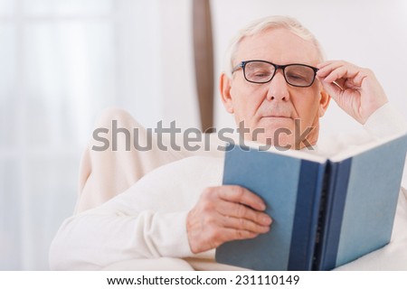 Enjoying time with favorite book. White hair senior man reading a book while sitting in chair at his apartment